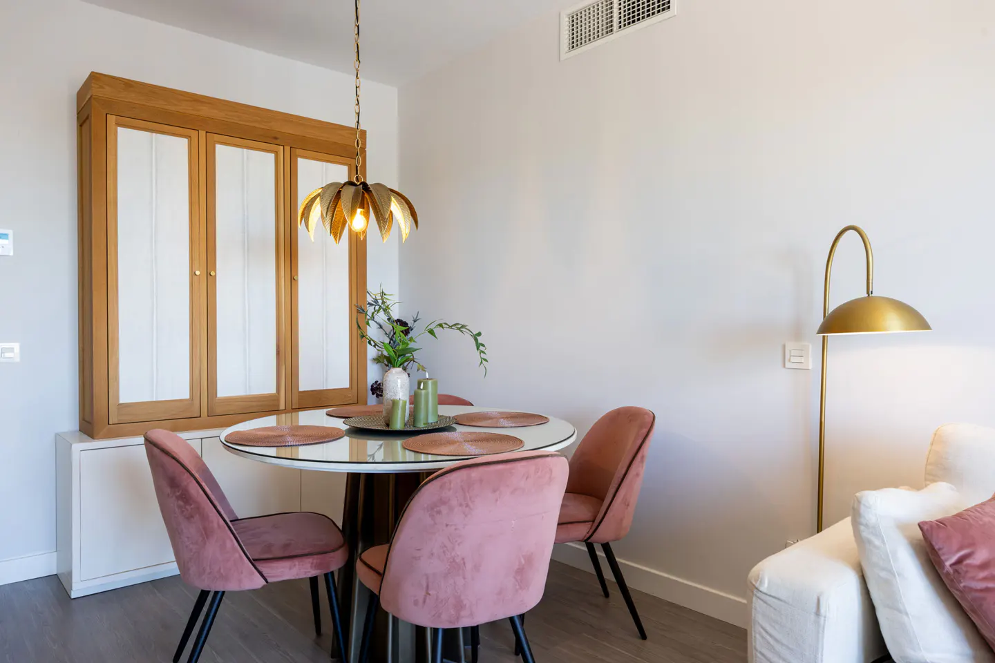 Dining area with round glass table, pink velvet chairs, and gold accents. A wooden cabinet is in the background.