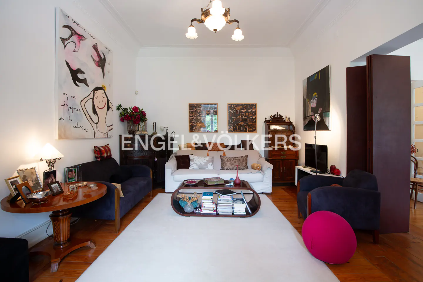 Bright living room with white walls, hardwood floors, and a large white rug. Sofas, chairs, art, and a coffee table with books furnish the space.