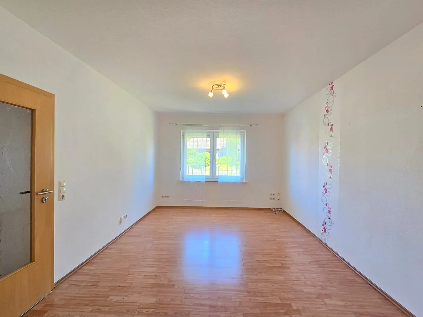 Empty room with light wood flooring, white walls, a window with sheer curtains, and a decorative floral wall accent.