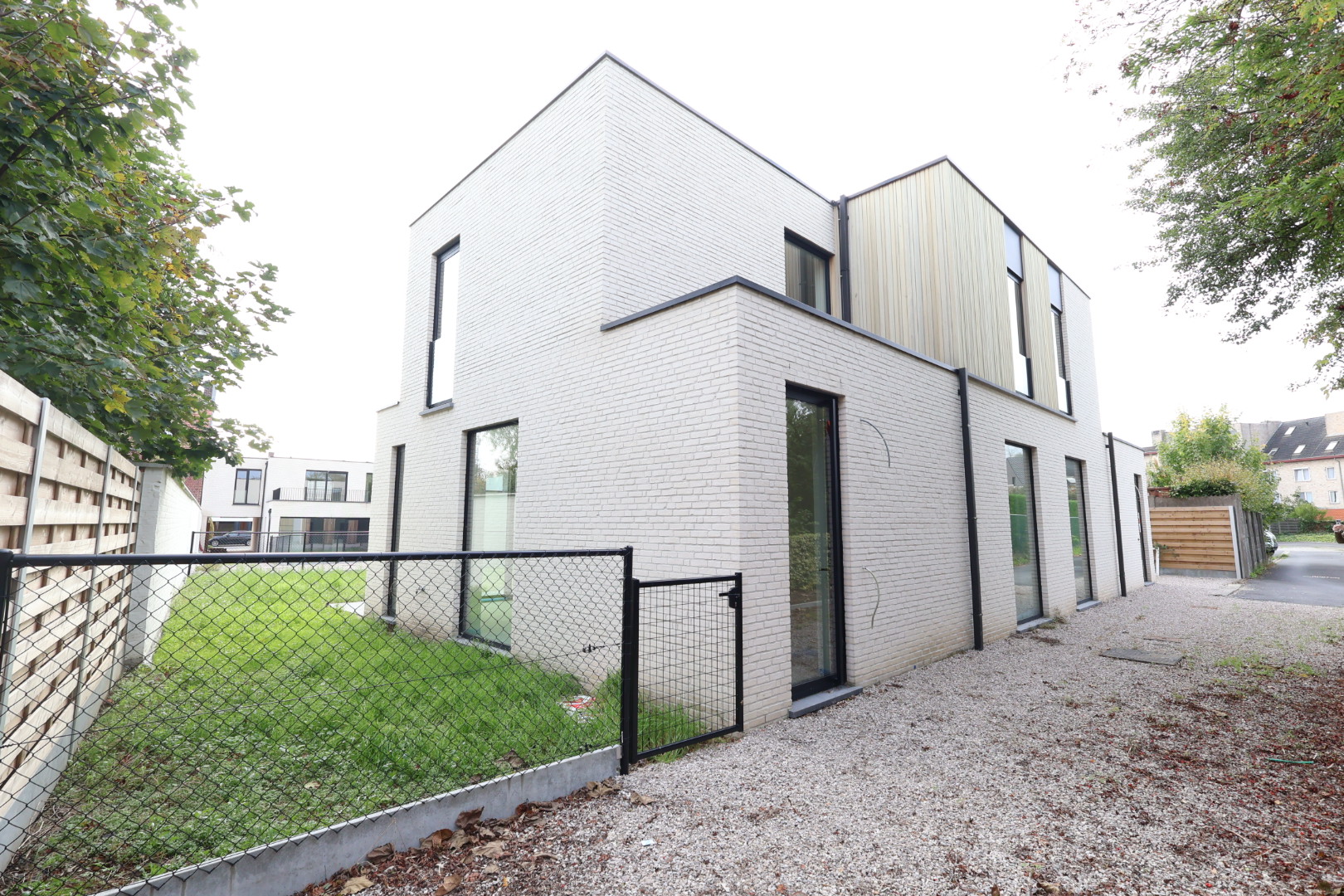 Modern two-story house with light brick and wood siding, large windows, a gravel driveway, and a fenced-in grassy yard. [AI-generated text]
