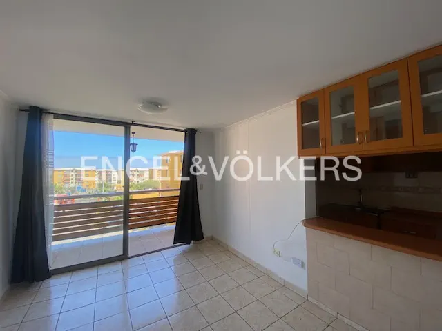 Bright apartment interior with balcony. Black curtains frame sliding glass doors to a balcony with a wooden railing. Kitchen cabinets are visible.
