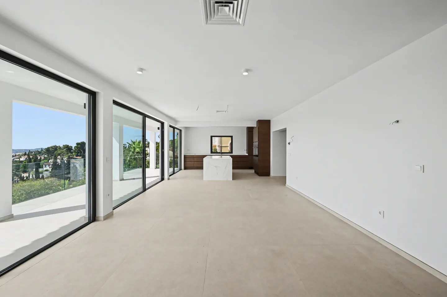 Bright, empty room with white walls, beige floor, and black-framed sliding glass doors. A marble island and dark wood cabinets are in the kitchen area.