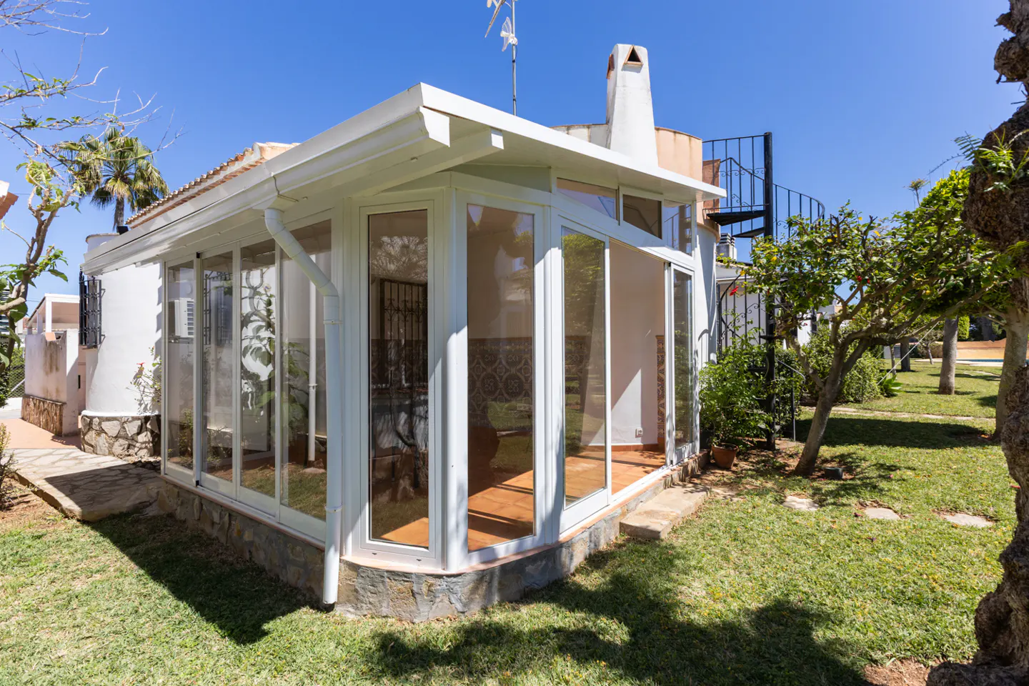 Exterior view of a white sunroom with glass walls, a spiral staircase, and a chimney, surrounded by green grass and trees under a blue sky.