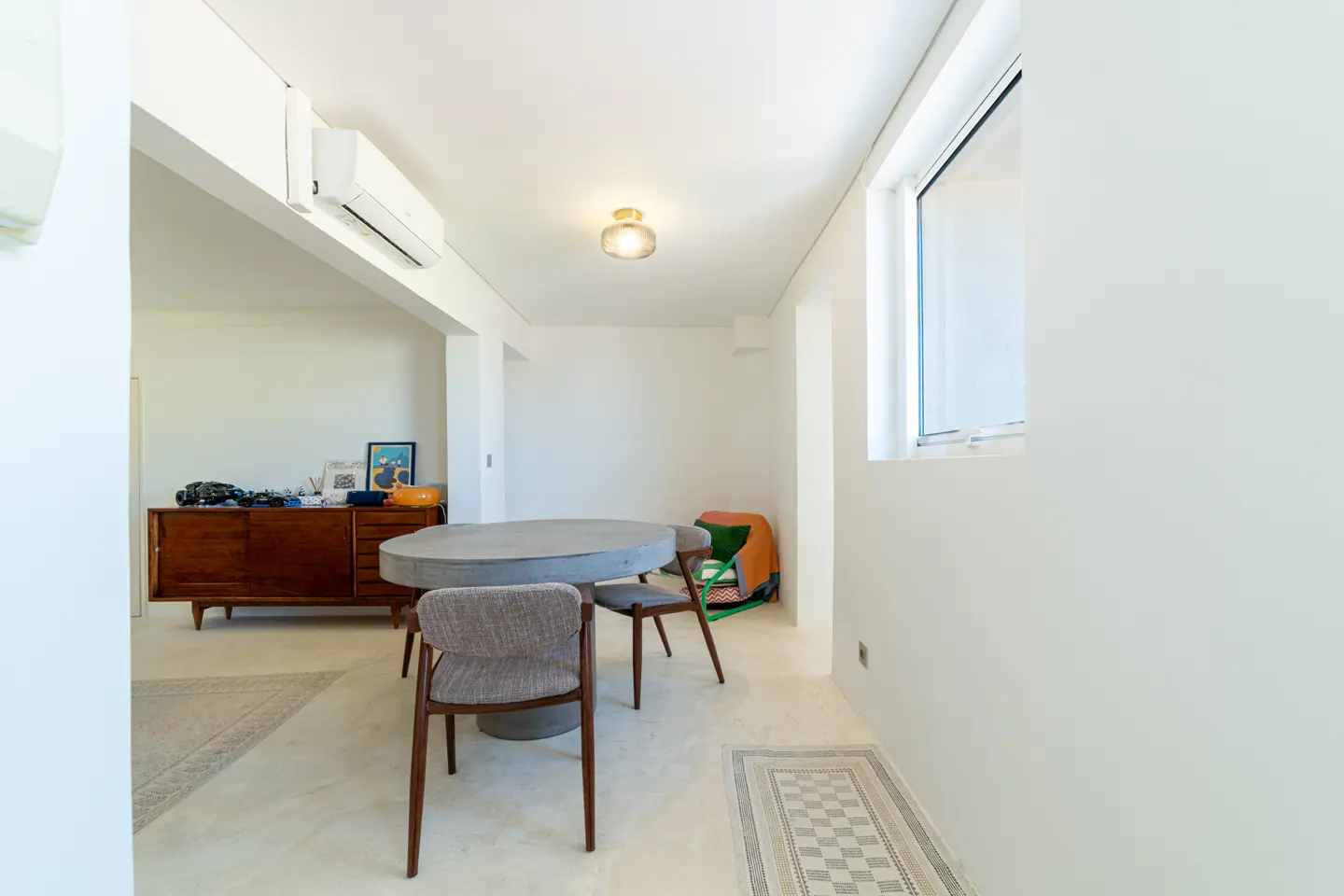 Bright, modern dining area with a round gray table, three chairs, and a wooden sideboard against white walls. A window lets in natural light.