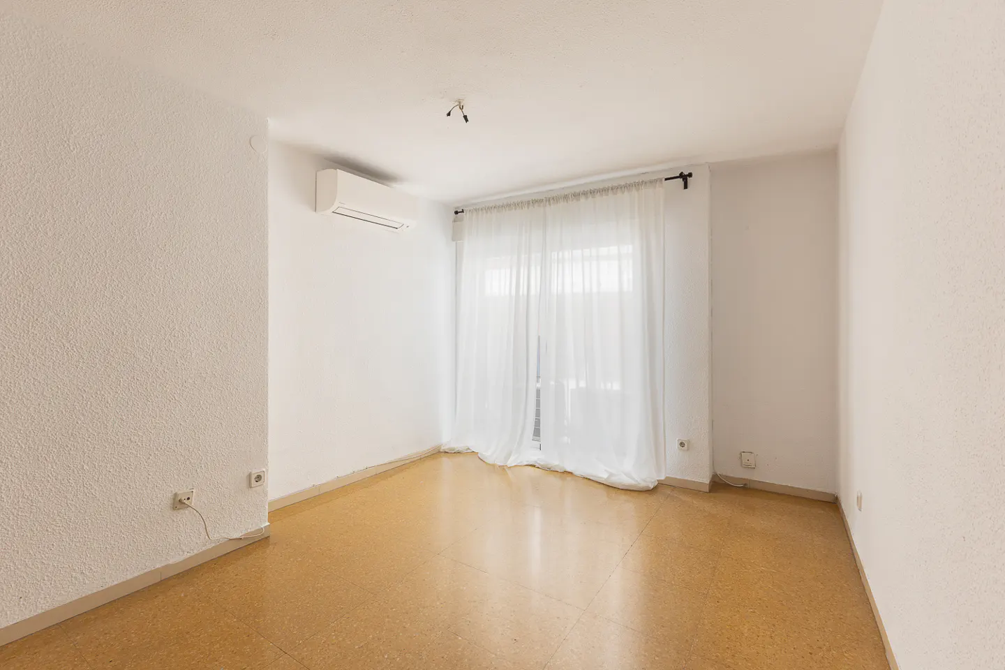 Empty room with white walls, tan tile flooring, an air conditioning unit, and sheer white curtains over a window.