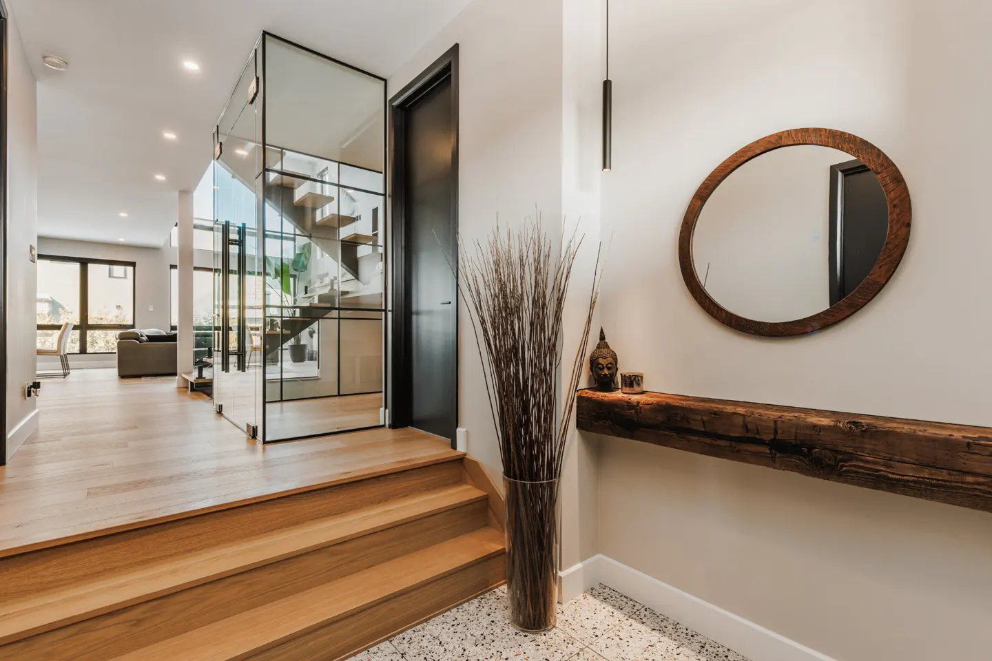 Modern entryway with wooden stairs, a glass-enclosed staircase, a round wooden mirror, and a rustic floating shelf.