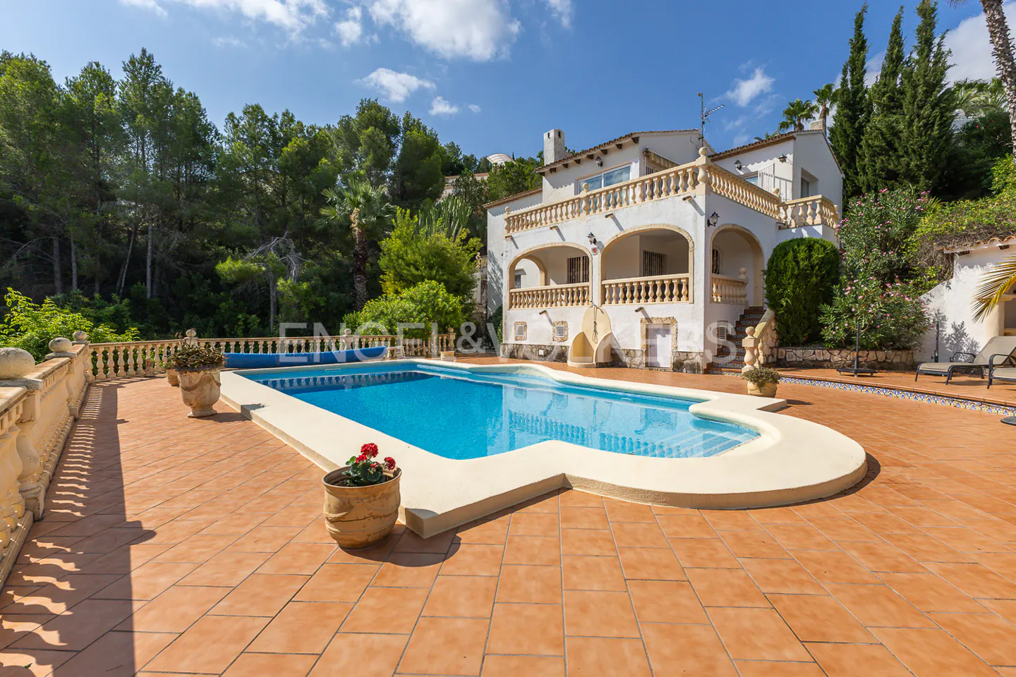A sunny view of a white two-story house with a blue swimming pool and terracotta patio. Trees and blue sky in the background.