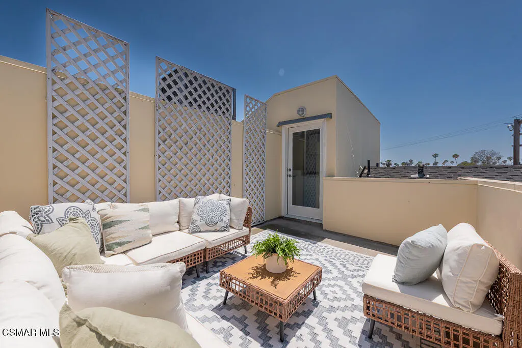 Outdoor patio with wicker furniture, white cushions, and a geometric rug. Lattice screens provide privacy. A door leads into the building.