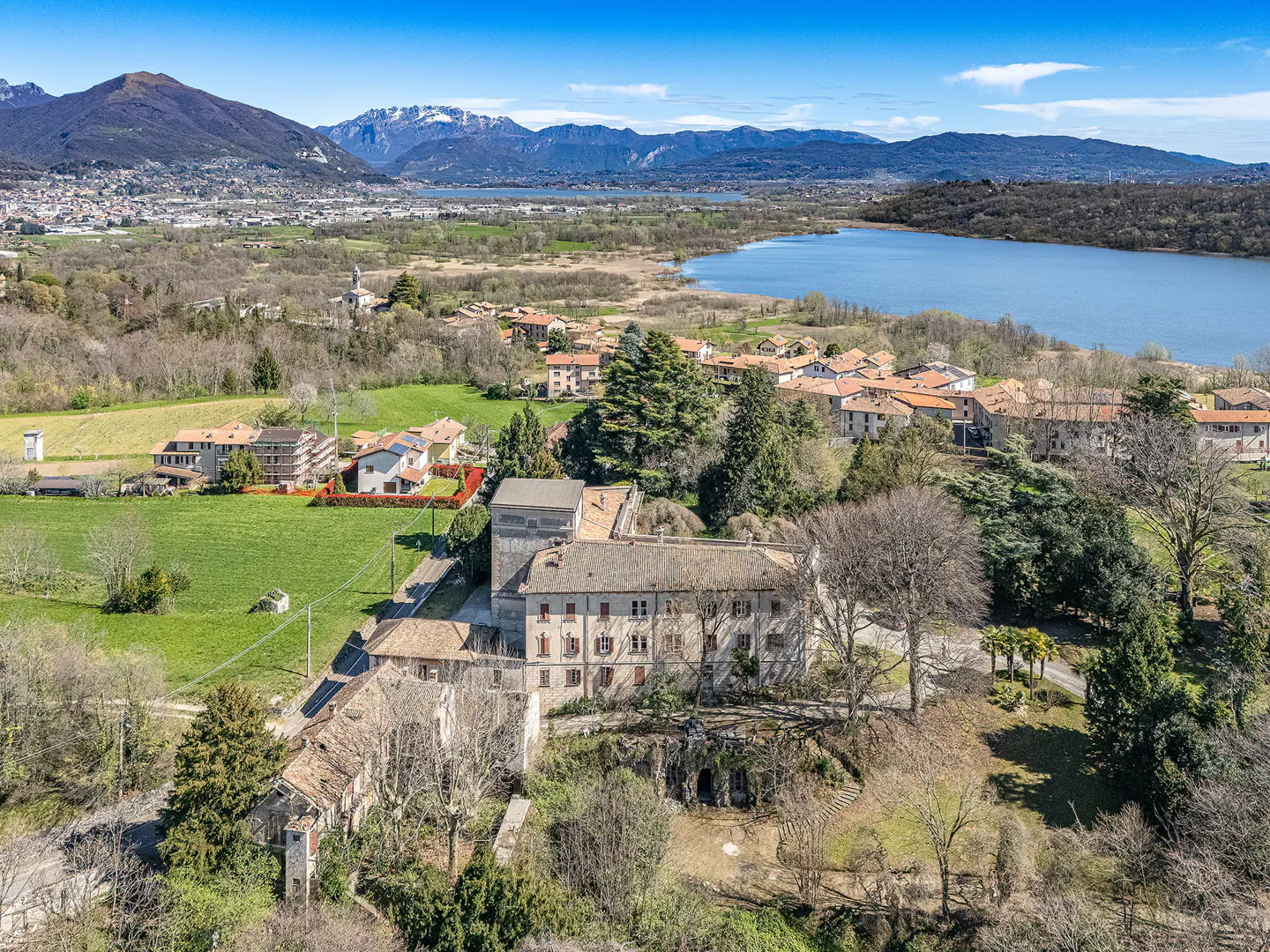 Aerial view of a large, old stone building surrounded by trees, with a lake and mountains in the background.