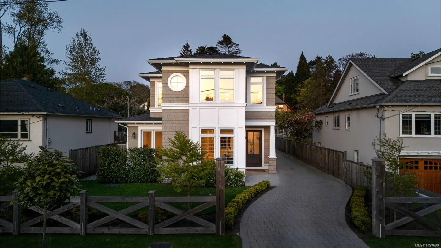 Two-story house with gray shingles, white trim, and a dark roof. A gray brick driveway leads to the front door. A wooden fence surrounds the front yard.
