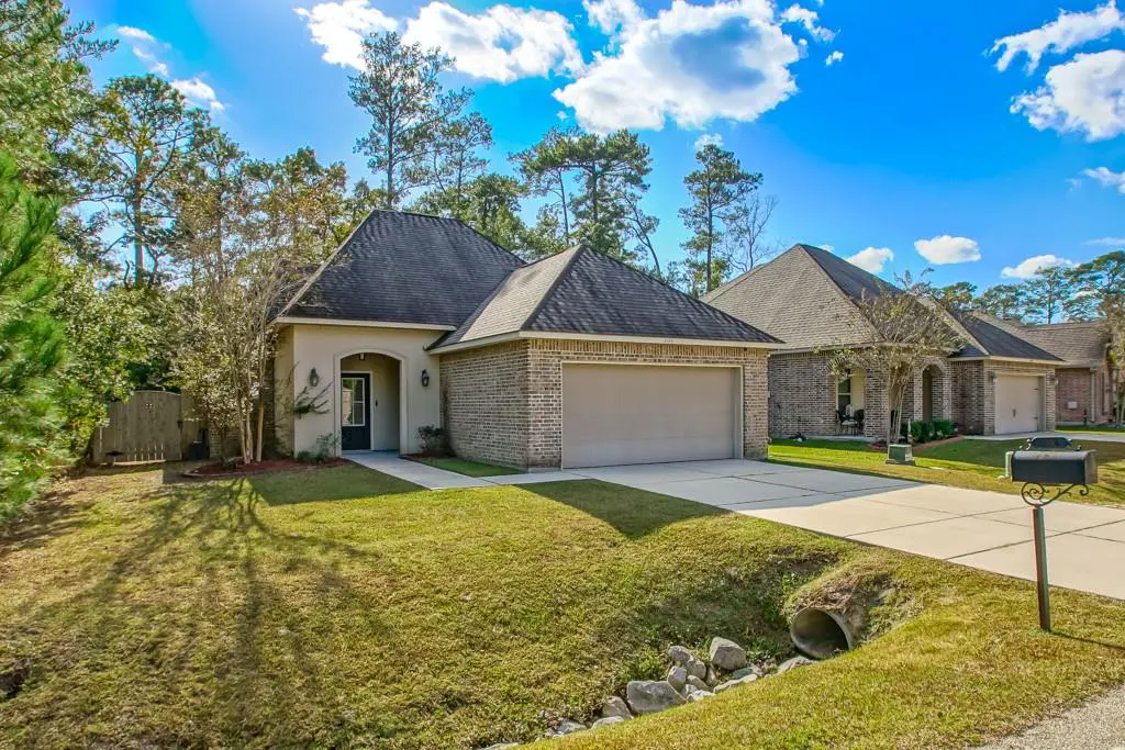 A single-story brick house with a dark roof, green lawn, and blue sky with white clouds.