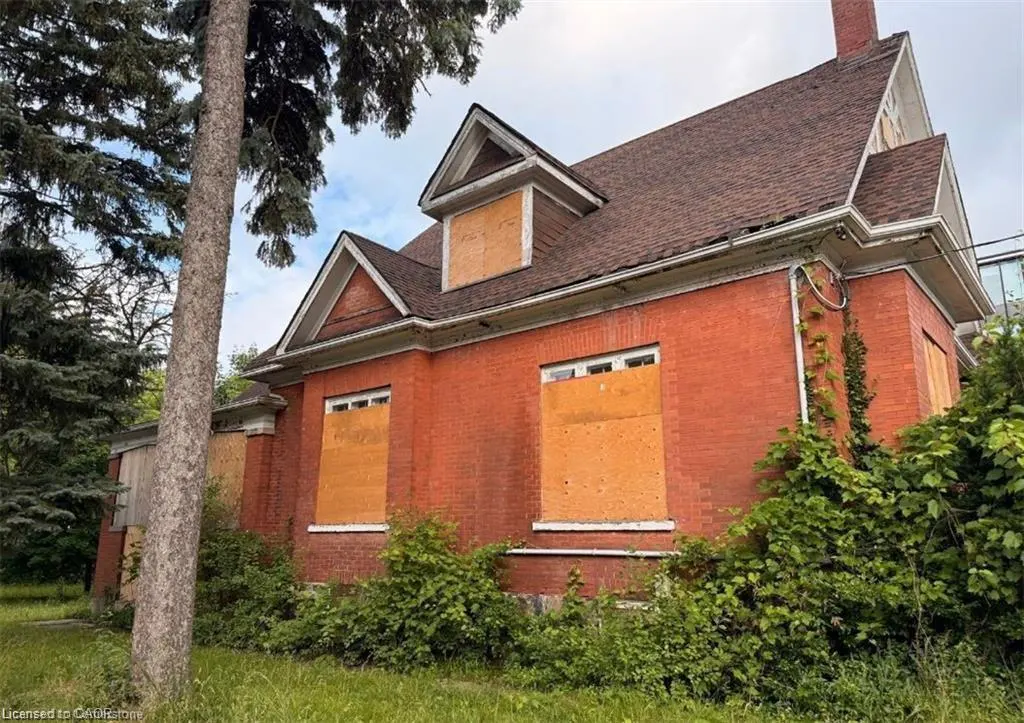 Exterior view of a red brick house with boarded-up windows and a brown shingled roof, surrounded by overgrown greenery.