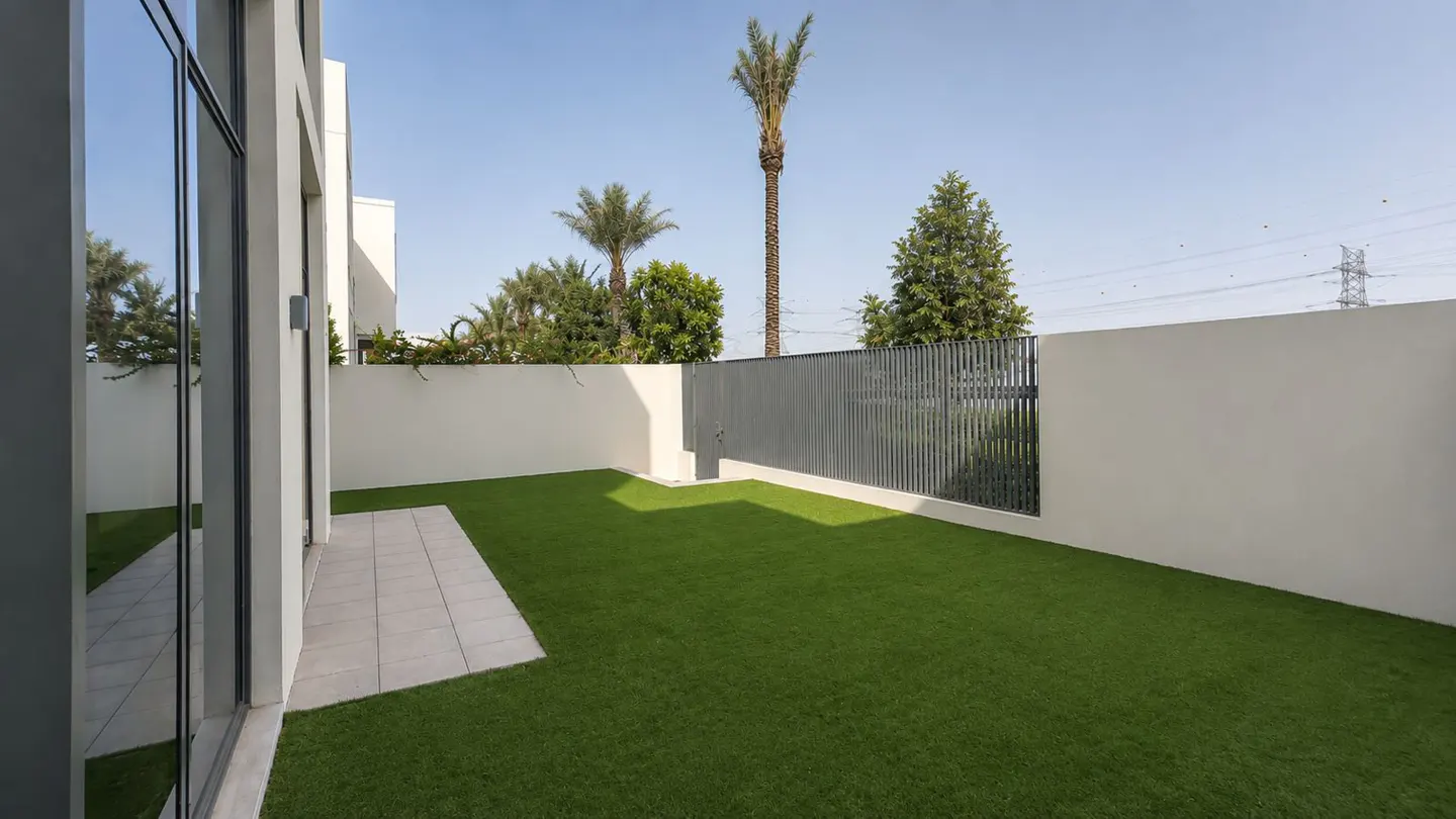 A modern backyard with green grass, a gray fence, and white walls under a blue sky. Palm trees are visible in the background.