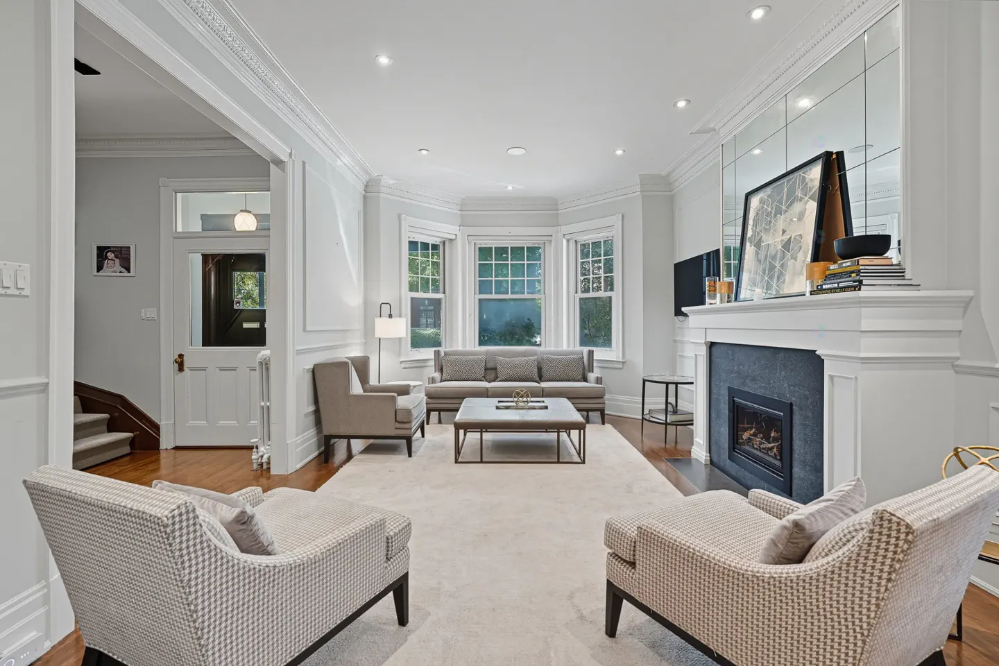 Bright living room with white walls, fireplace, and bay window. Two patterned chairs face a gray sofa and coffee table on a cream rug.