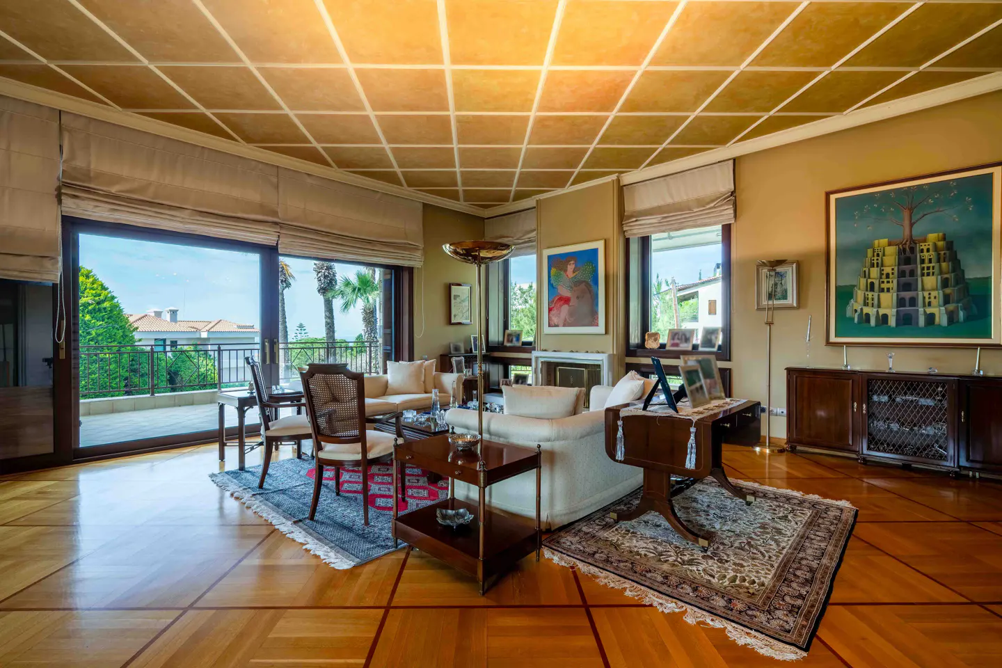 A living room with wood floors, beige walls, and a coffered ceiling. There are sofas, chairs, tables, and artwork. A balcony is visible through large windows.