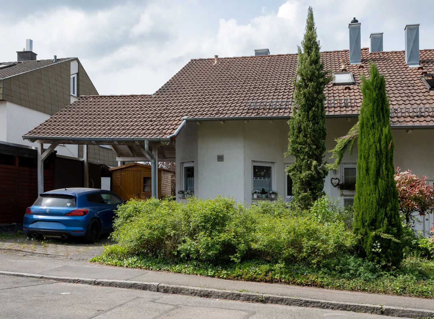 A one-story house with a brown tile roof, white walls, and a blue car parked under a carport.