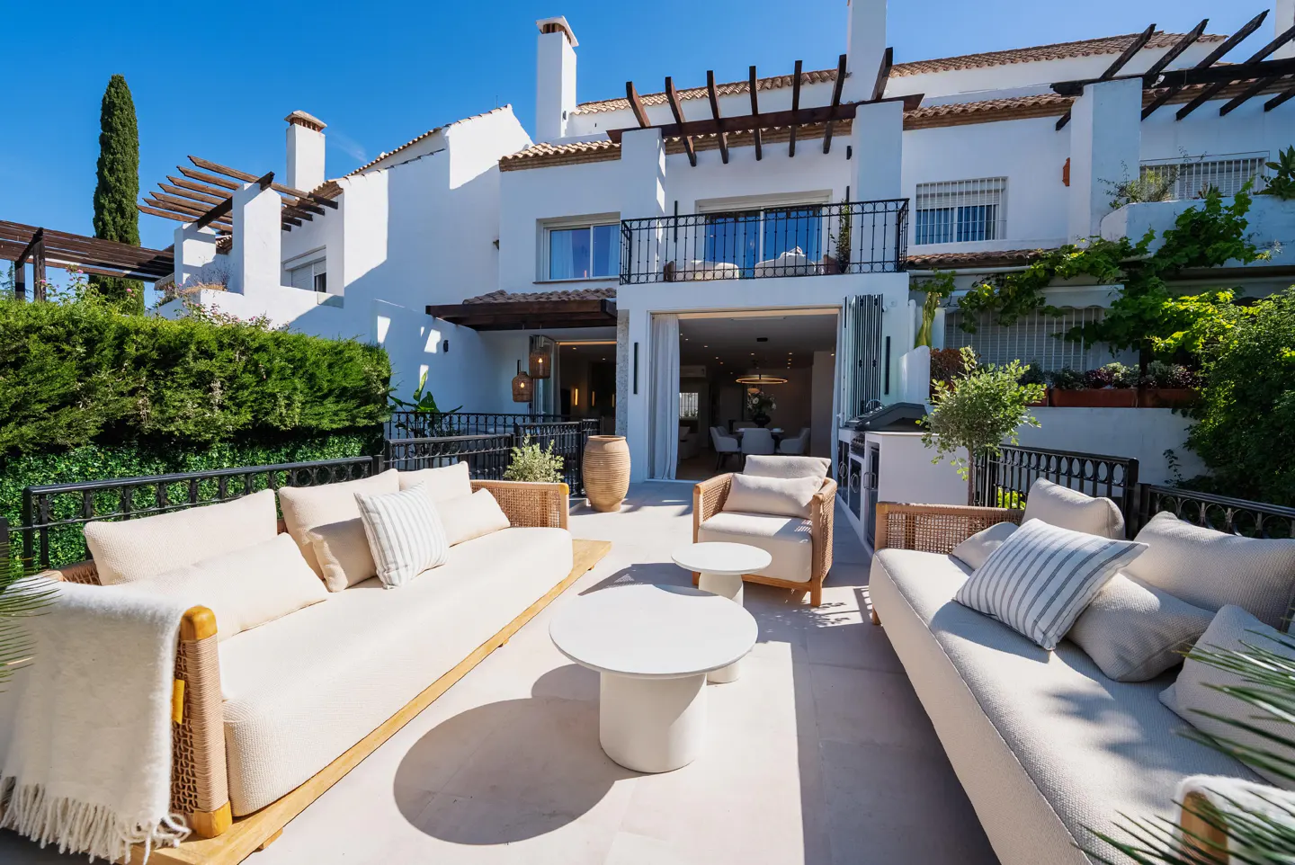 Outdoor patio with white sofas, chairs, and tables. White building with dark wood pergola in the background. Green hedges and blue sky.
