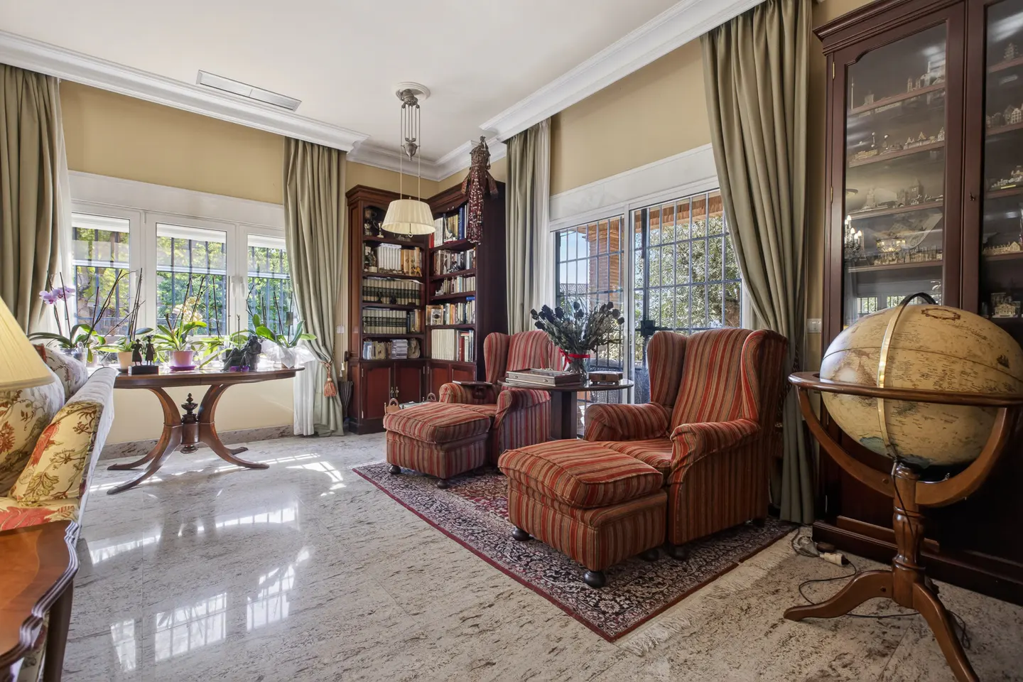 A living room with striped armchairs, a globe, and bookshelves. The room has beige walls and curtains.
