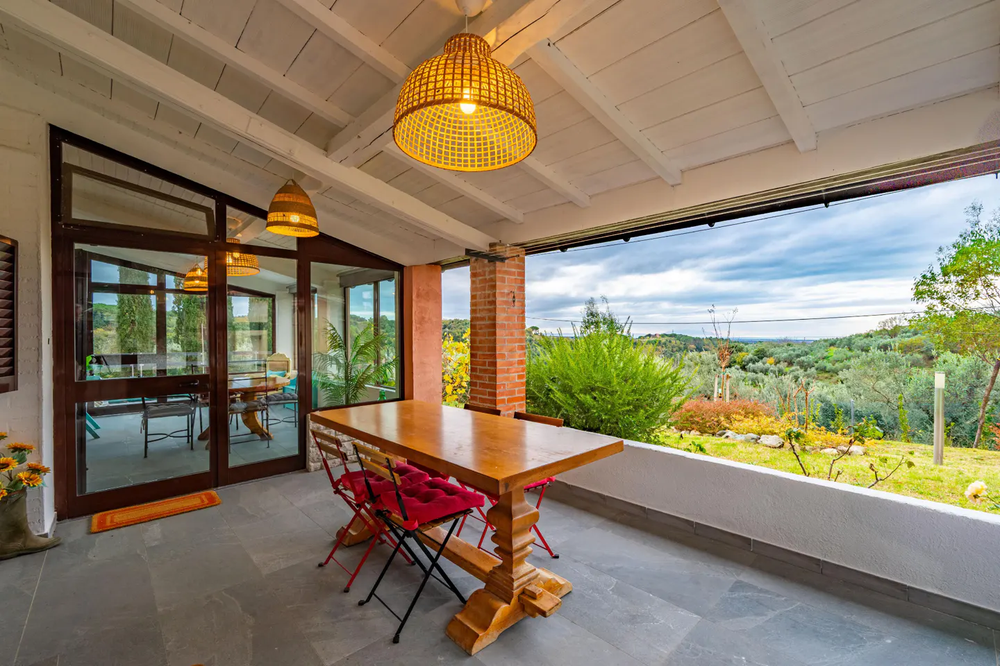 Covered patio with a wood table, red chairs, and woven pendant lights. A view of green hills is visible.
