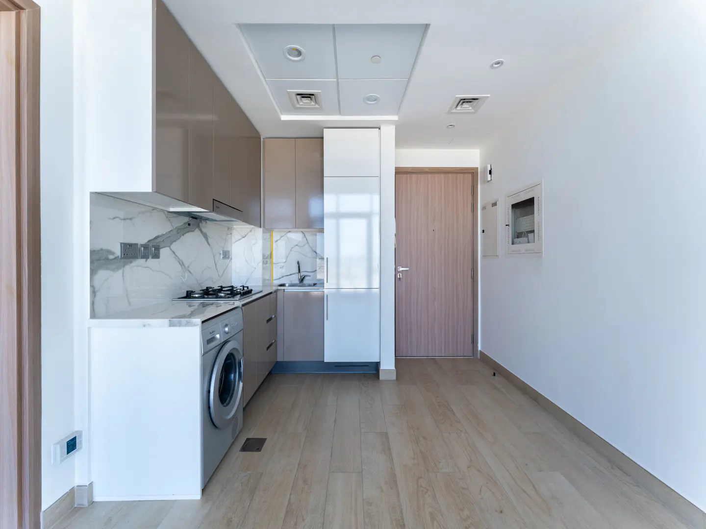 A modern kitchen with light wood floors, white walls, and taupe cabinets. A washing machine is built into the kitchen cabinets.