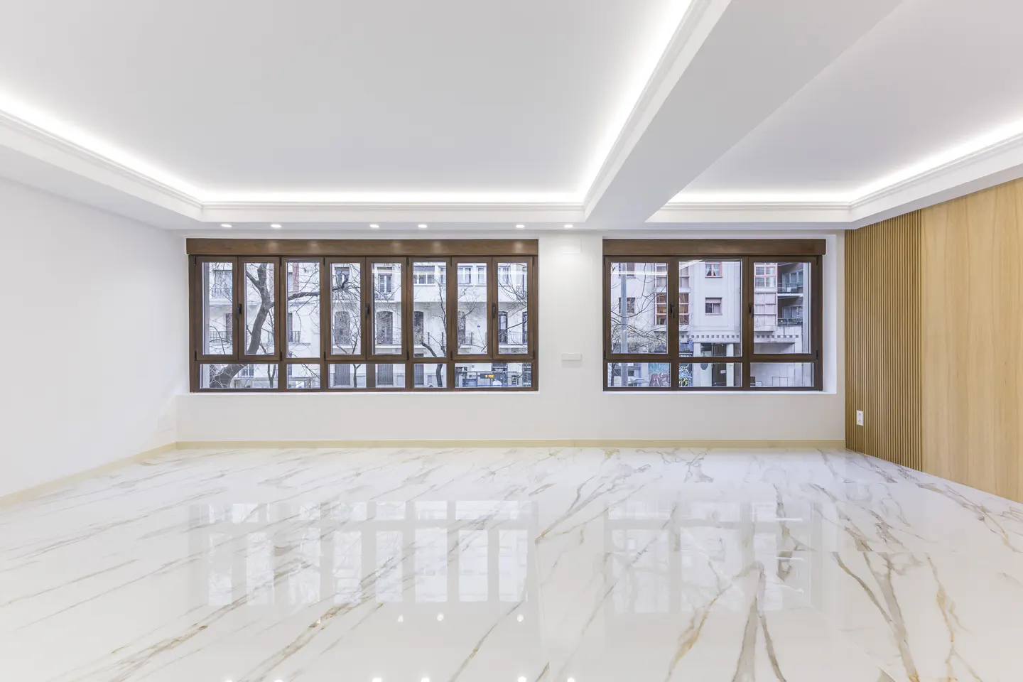 Bright, empty room with white marble floors, white walls, and two windows with brown frames. Outside, buildings and trees are visible.