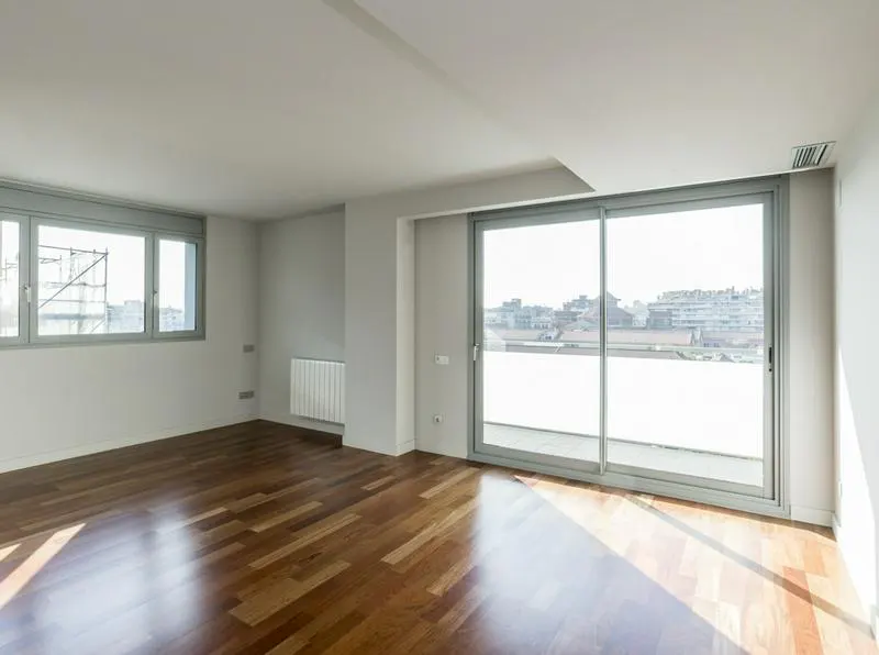 Bright, empty room with hardwood floors, white walls, and large sliding glass doors to a balcony with a city view.