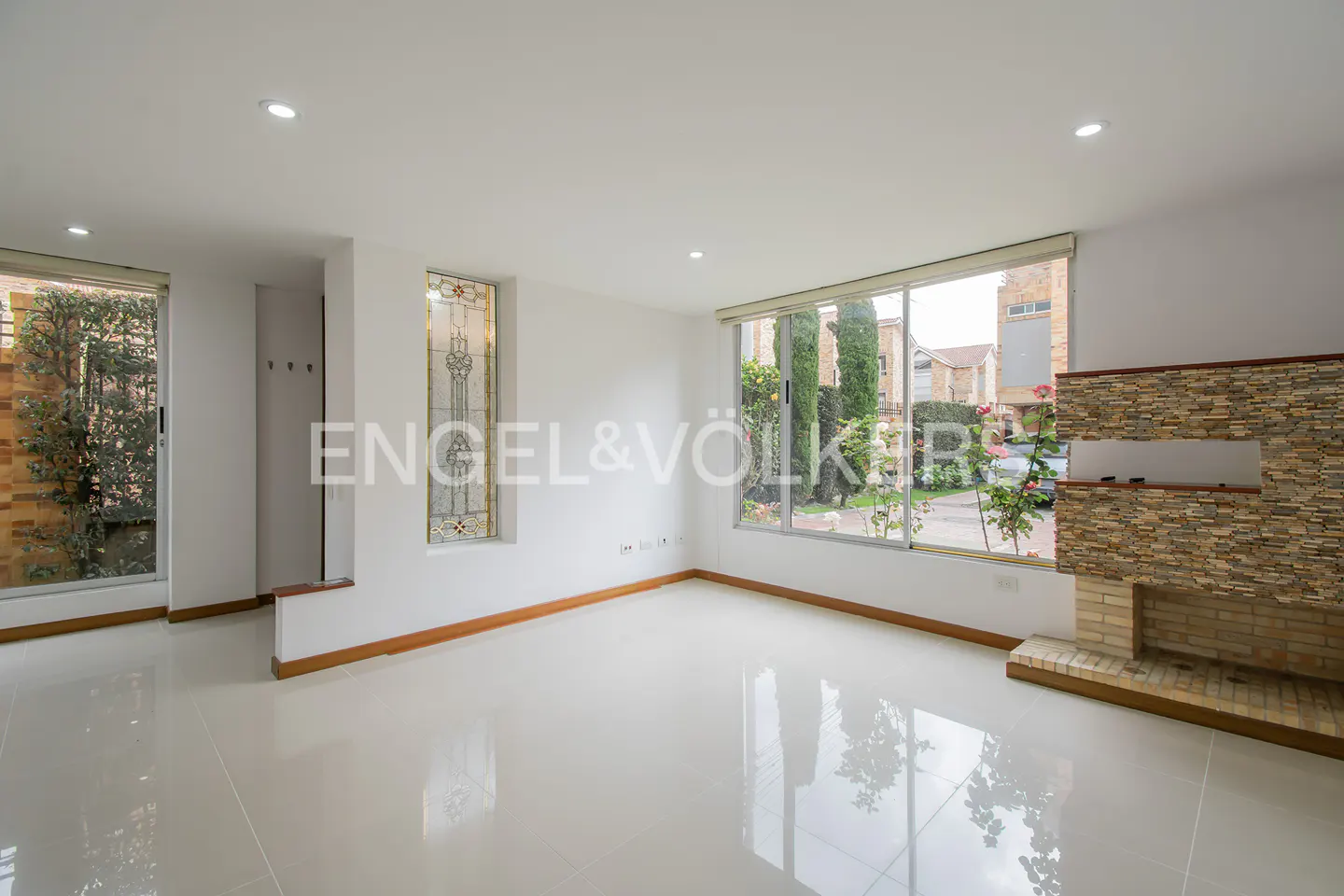 Bright, empty living room with white walls and tile floors. Large windows offer views of greenery and a brick fireplace adds texture.