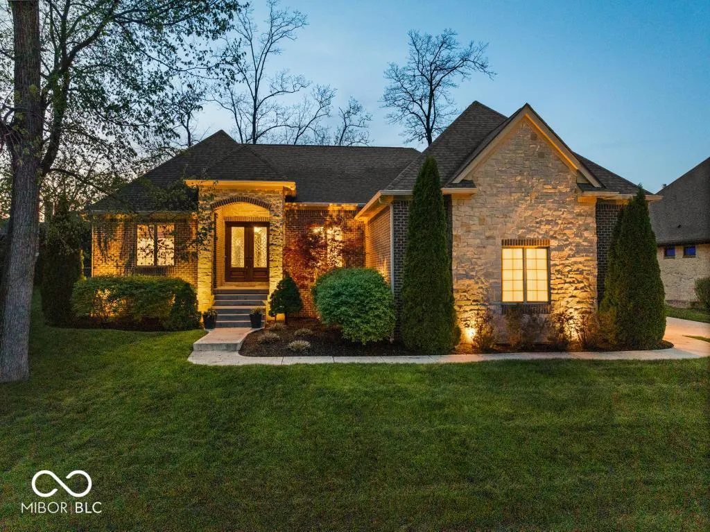 A single-story brick and stone house with a dark roof, green lawn, and illuminated windows at dusk.