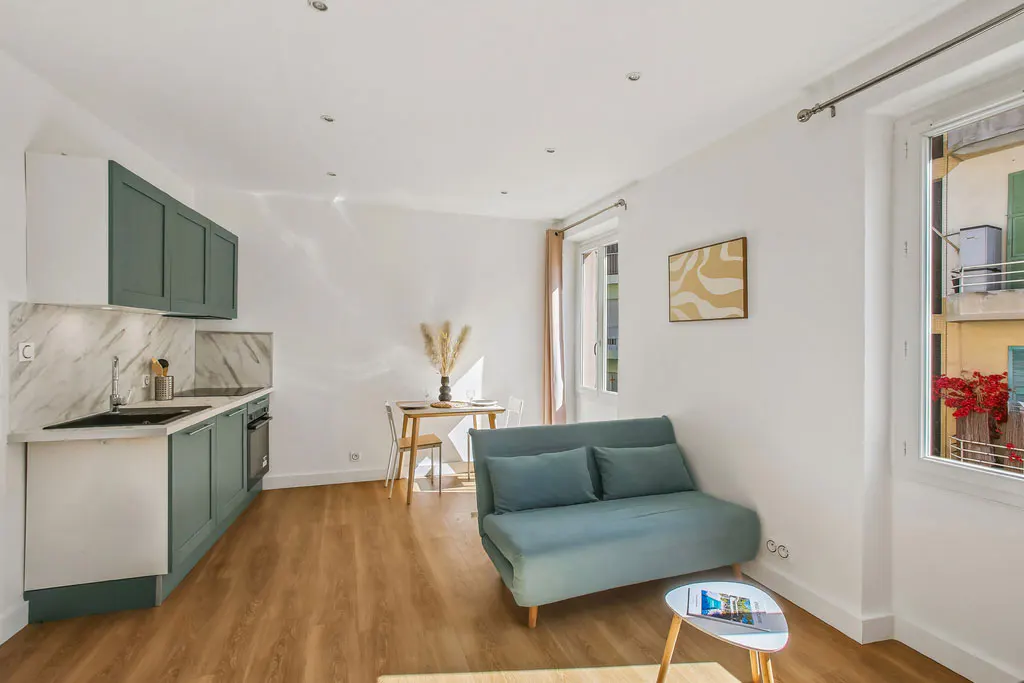 Bright studio apartment with wood floors, white walls, and green cabinets. A blue sofa sits near a window with red flowers outside.