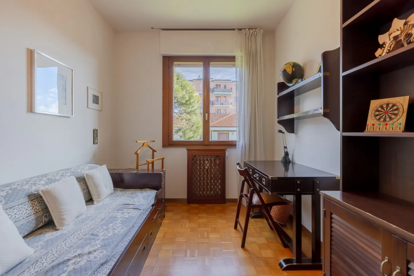 A cozy room with a daybed, desk, and dark wood shelving unit. A window with a view of buildings and a tree.