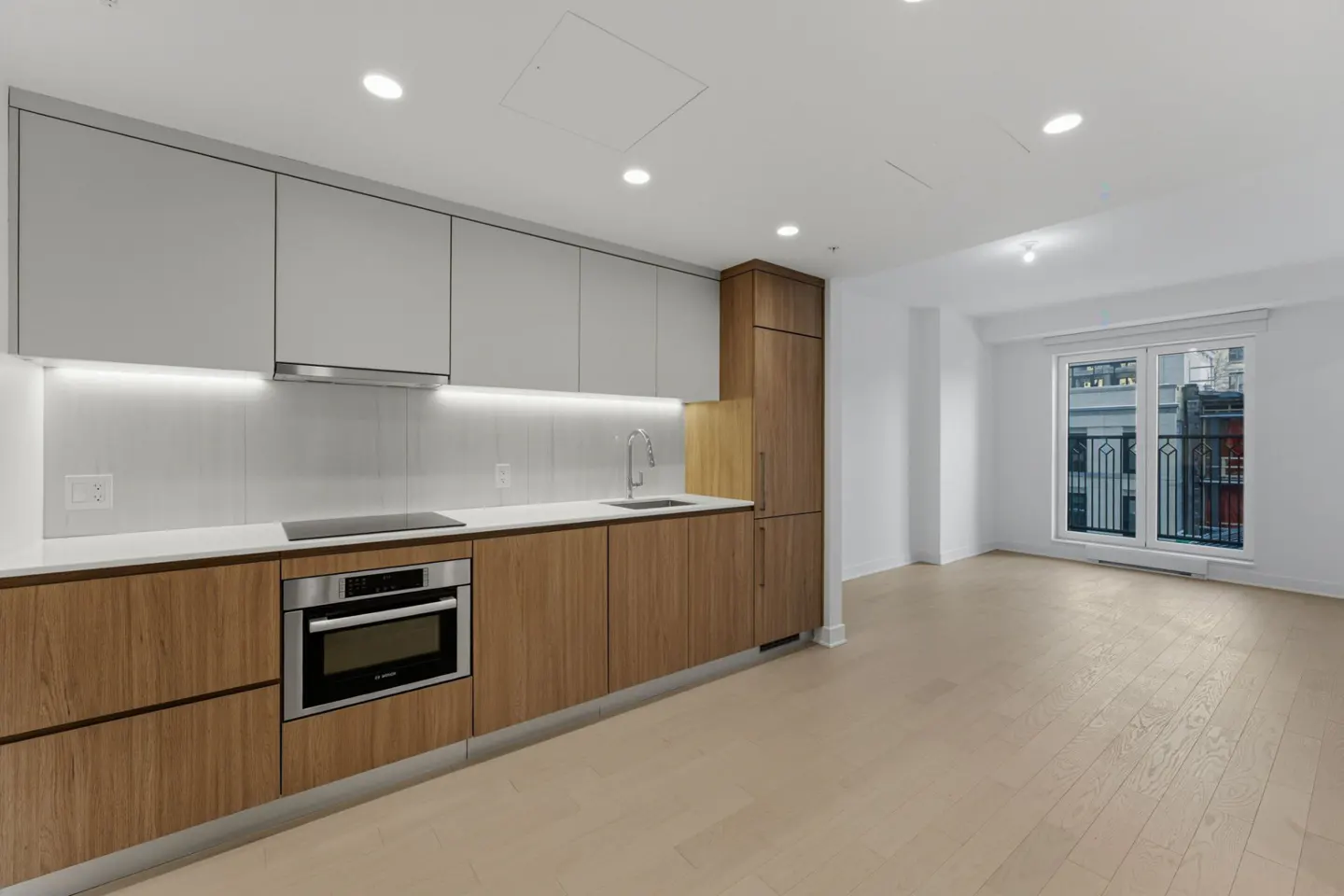 Modern kitchen with gray and wood cabinets, stainless steel oven, and white countertops. Light wood floors lead to a window with a city view.