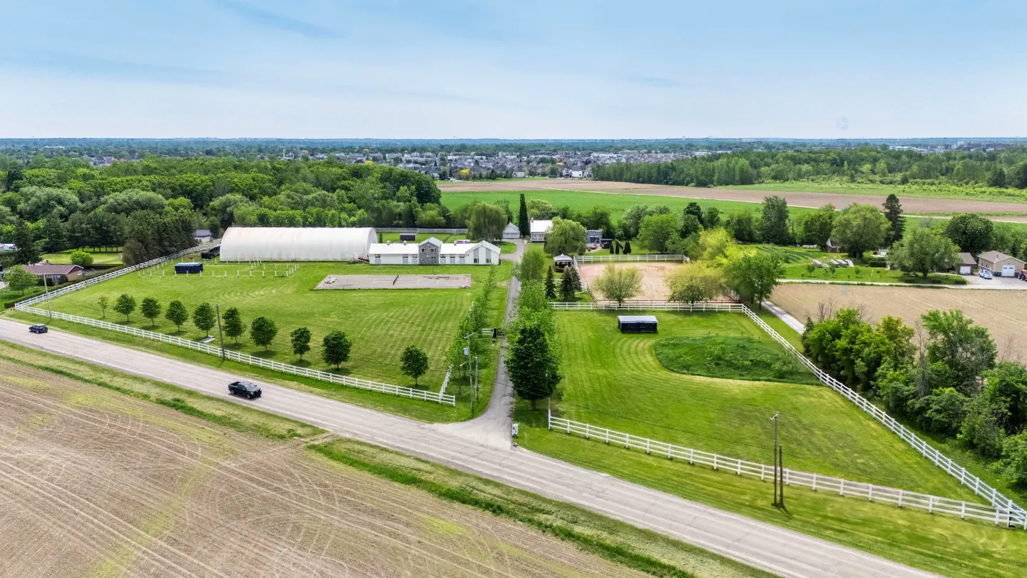 Aerial view of a rural property with a large white barn, fenced pastures, and green fields next to a paved road.