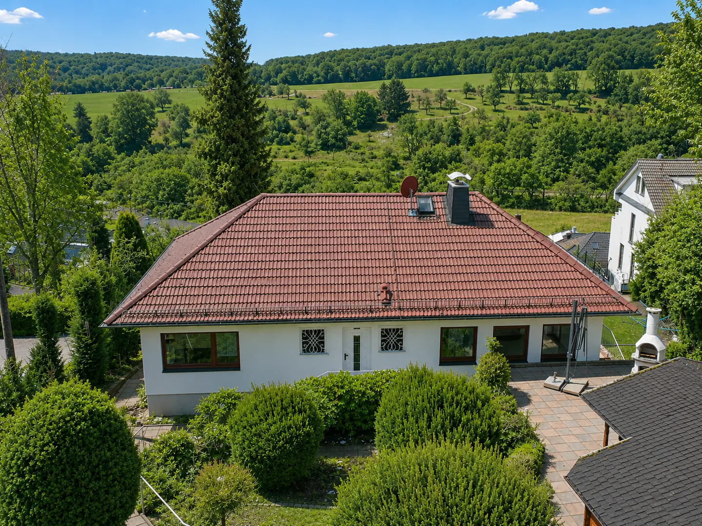 Aerial view of a white house with a red tile roof, surrounded by green trees and bushes, with a green field in the background.
