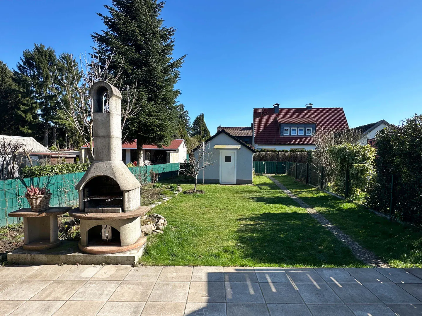 Backyard view with a stone barbecue, green lawn, and a small shed under a clear blue sky.