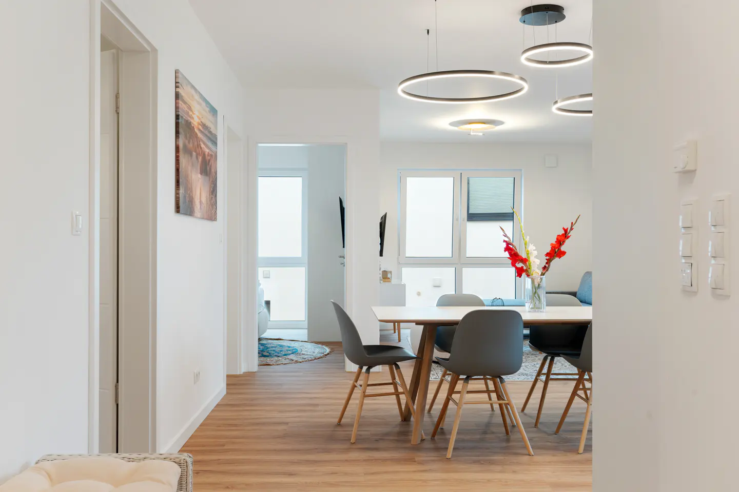 Bright, modern dining area with white walls, wood floors, and ring-shaped pendant lights. A white table with gray chairs sits near a window.