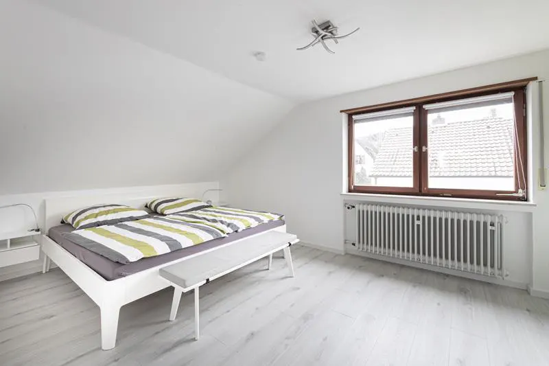 Bright bedroom with white walls, light wood floors, and a large window. A white bed with striped bedding and a bench sits in the center.