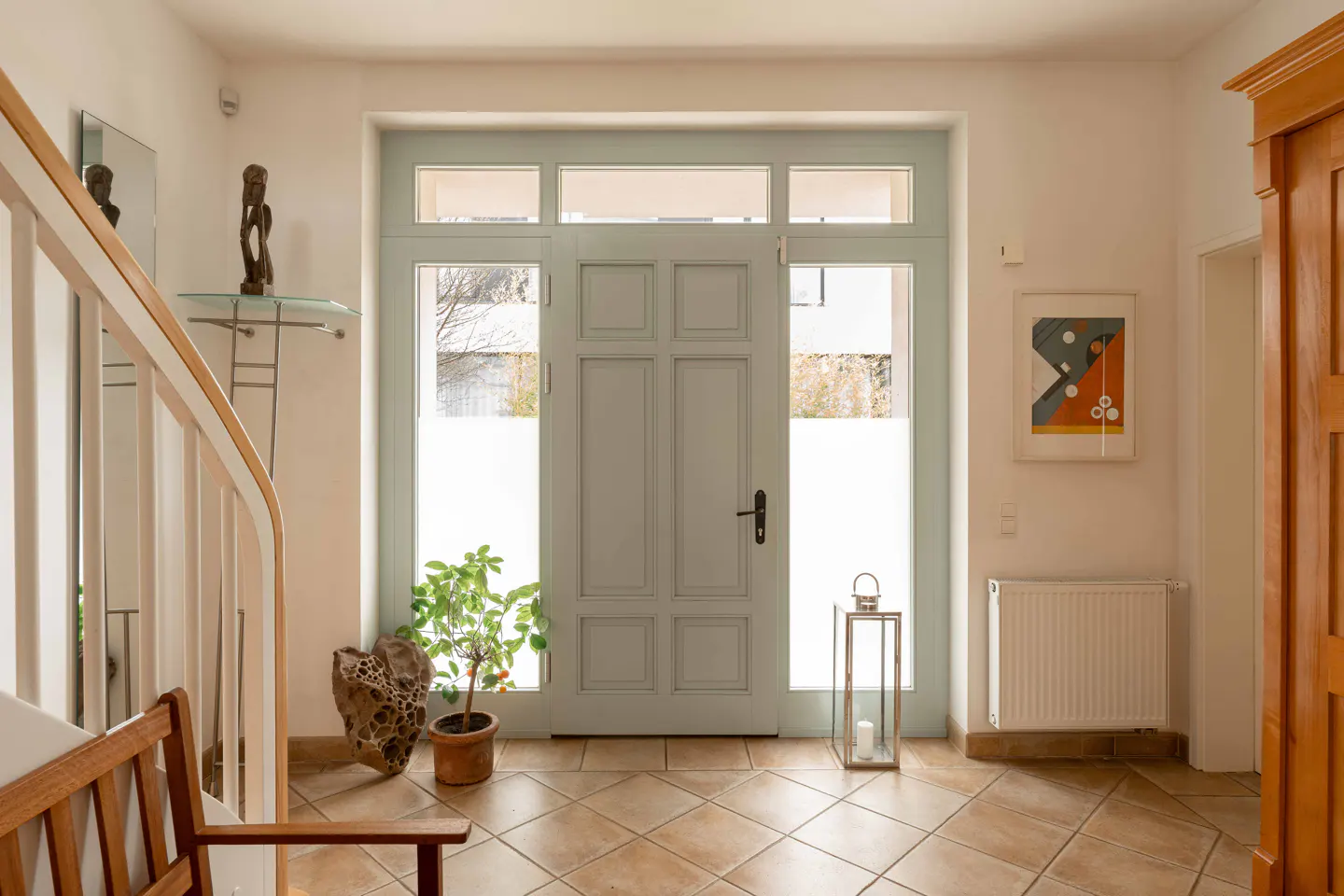 Bright entryway with a light blue door, tiled floor, a potted plant, a lantern, and a wooden staircase.