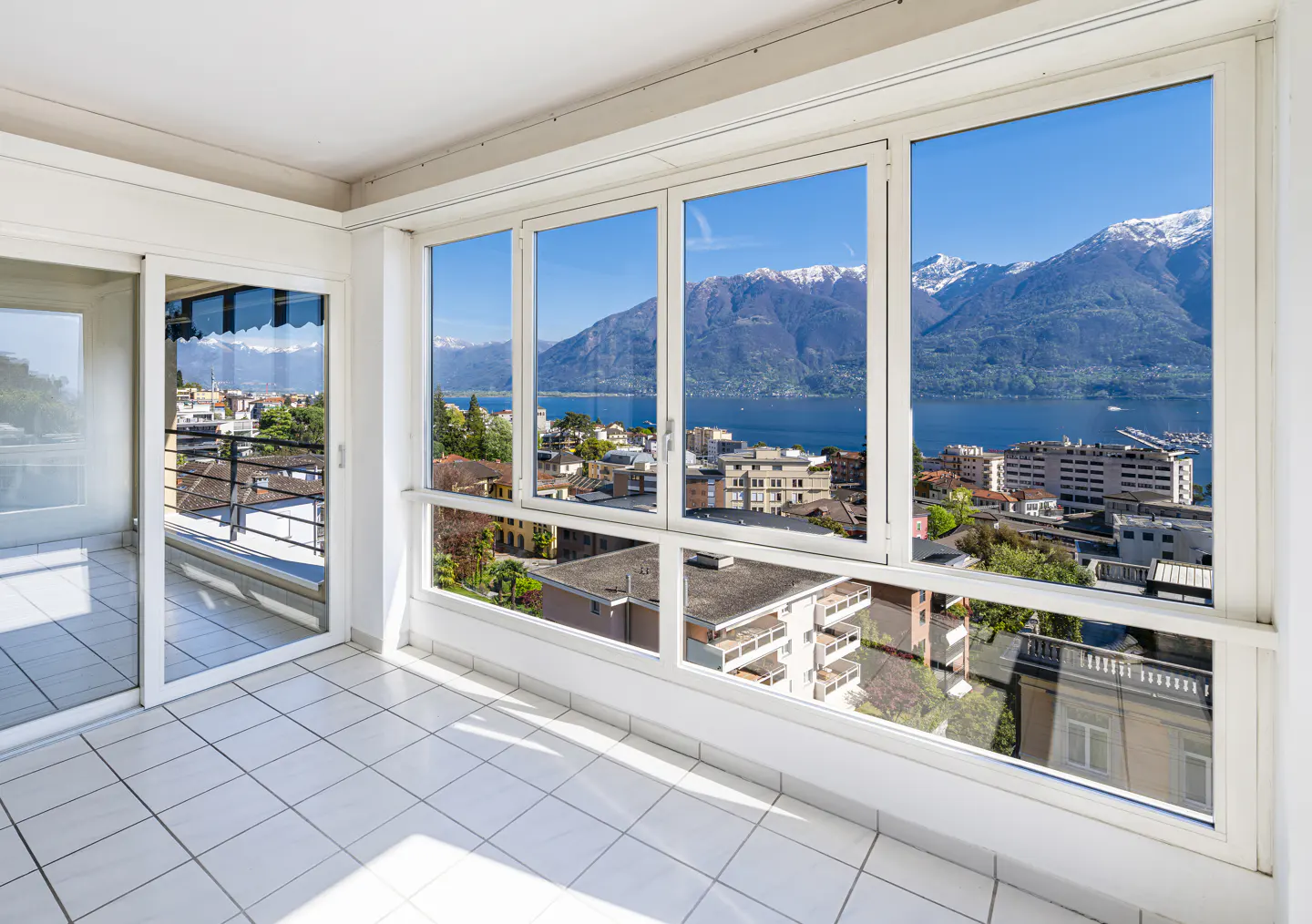 Bright sunroom with white tile floor and large windows. Outside, a city view with mountains and a blue lake.