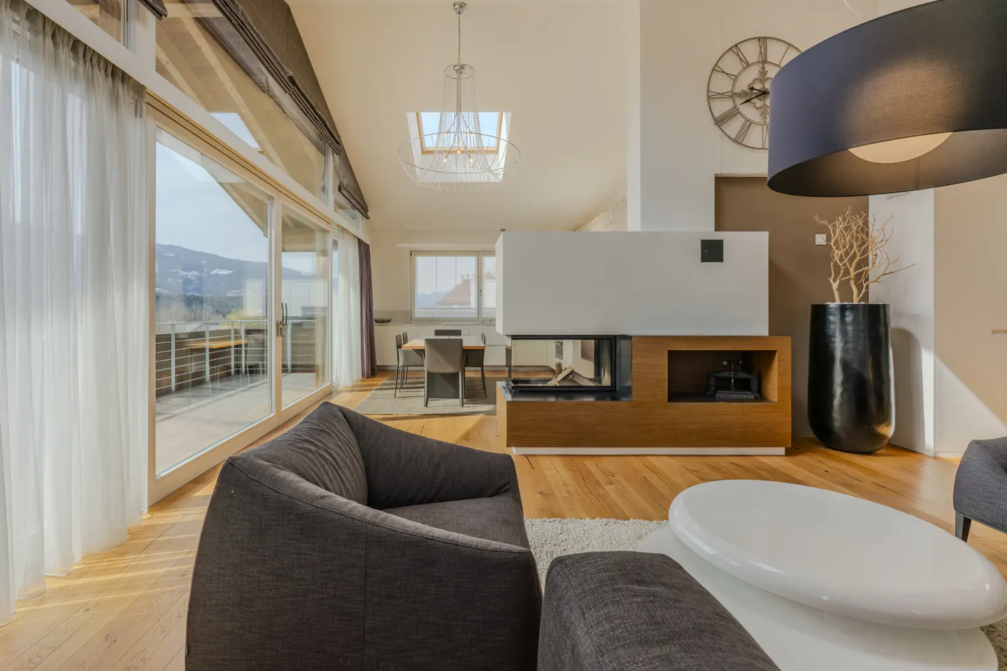 Bright living room with hardwood floors, a gray sofa, and a modern fireplace. Large windows offer a view of mountains.