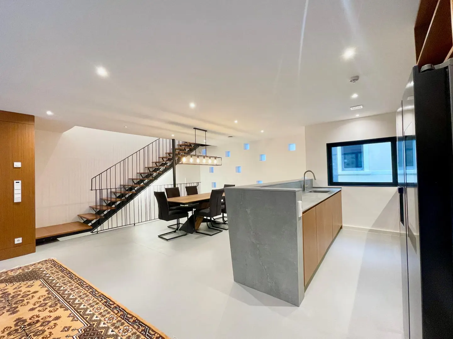 Open-concept living space with a staircase, dining table, and kitchen island. The walls are white with blue accents. A patterned rug is on the floor.