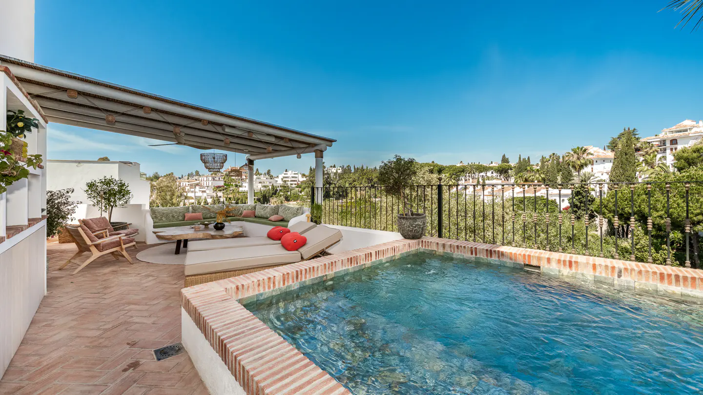 Rooftop terrace with a small pool, lounge area with a sofa, chairs, and a coffee table. The pool has brick trim. City view in the background.