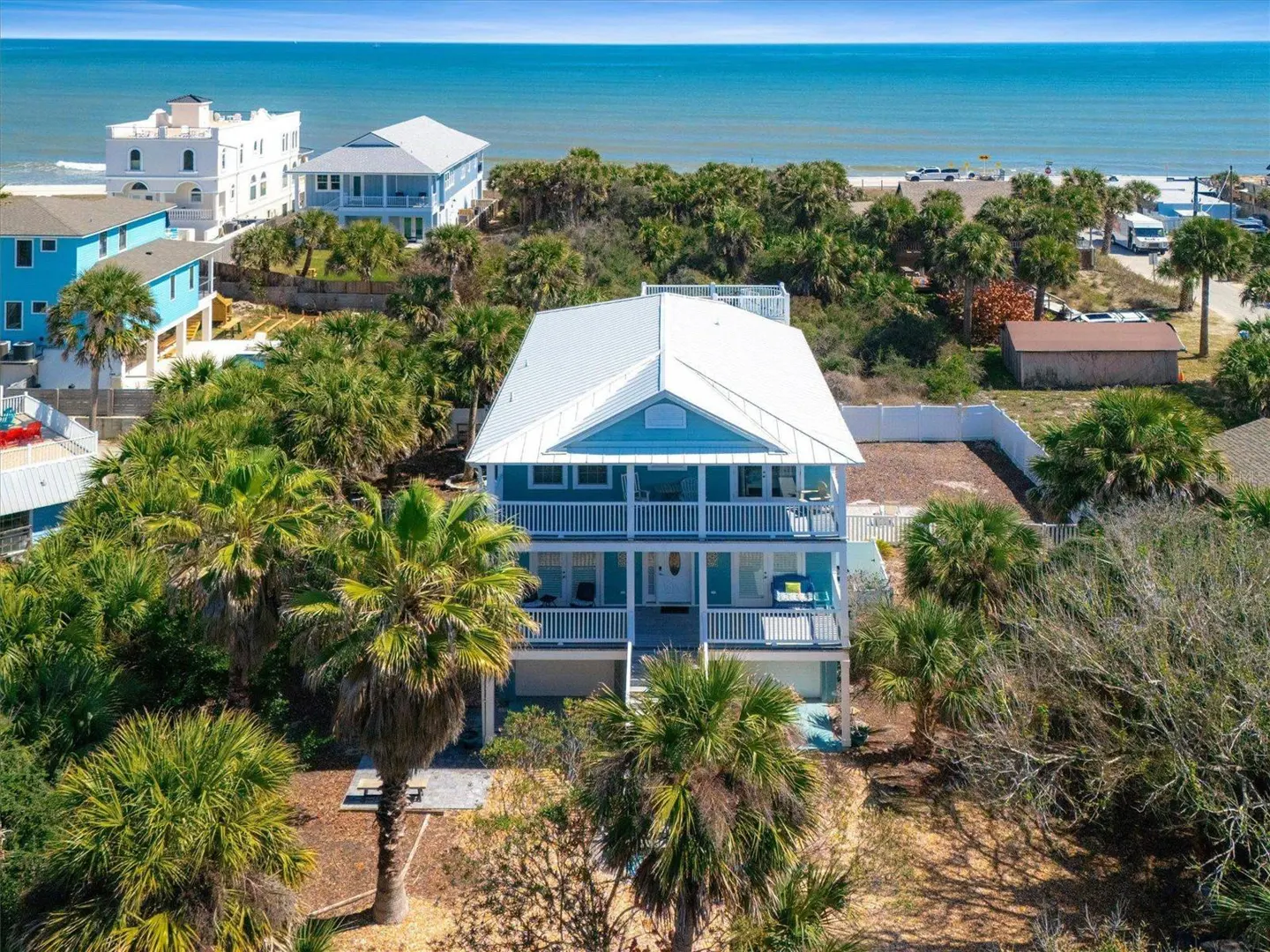 Aerial view of a three-story light blue beach house with white trim and a metal roof, surrounded by palm trees, with the ocean in the background.