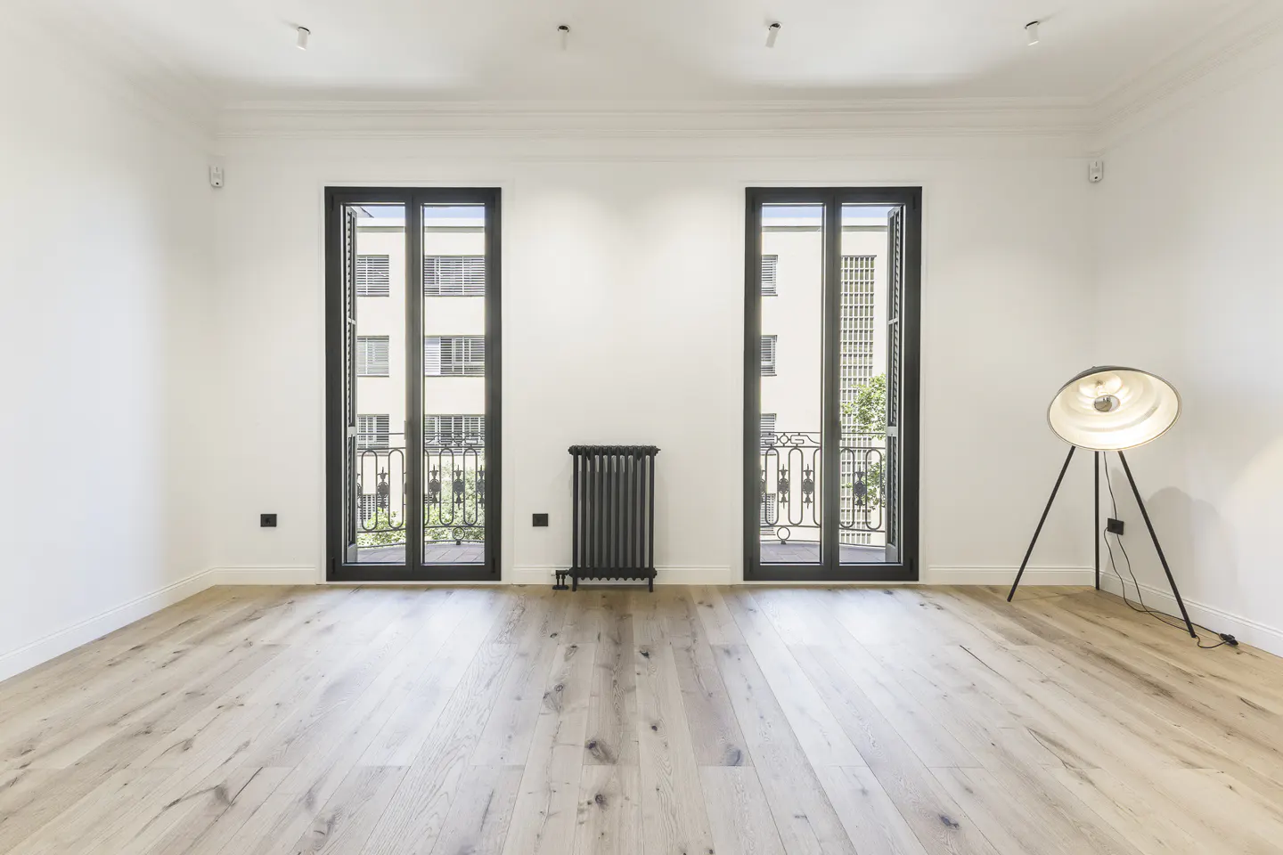 Bright, empty room with light wood floors, white walls, and black-framed windows with balconies. A black radiator and a tripod floor lamp are also visible.