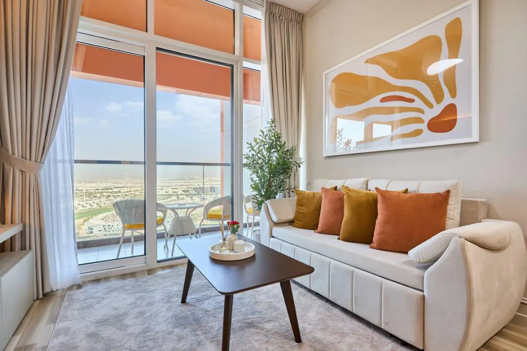 Living room with a white sofa, orange pillows, and a dark wood coffee table. Balcony with city view through large windows. Abstract art on the wall.