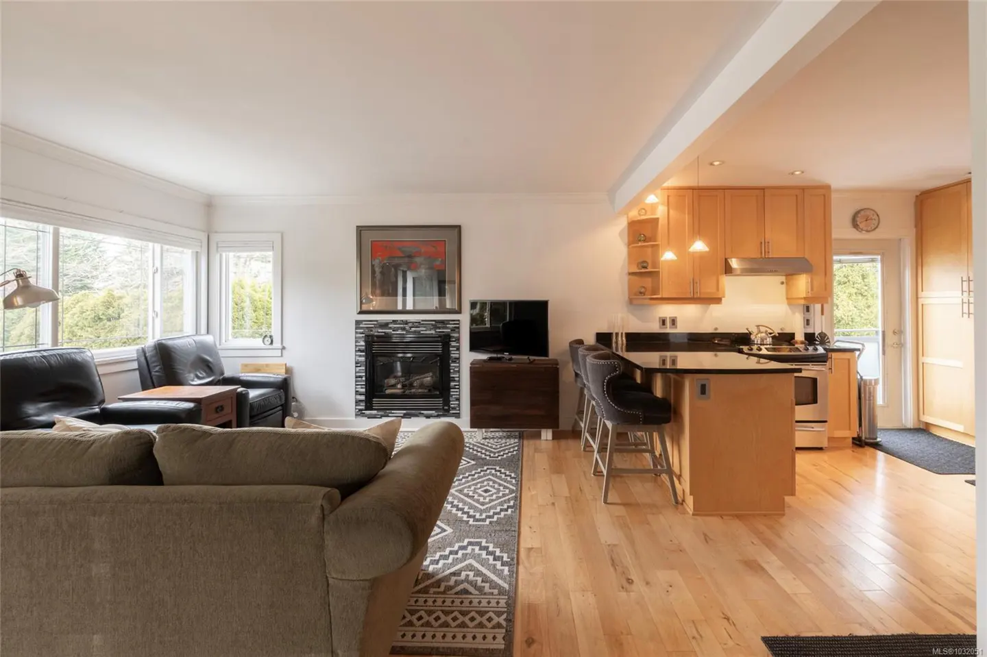 Open-concept living space with a kitchen, fireplace, and seating area. Hardwood floors and natural light fill the room.