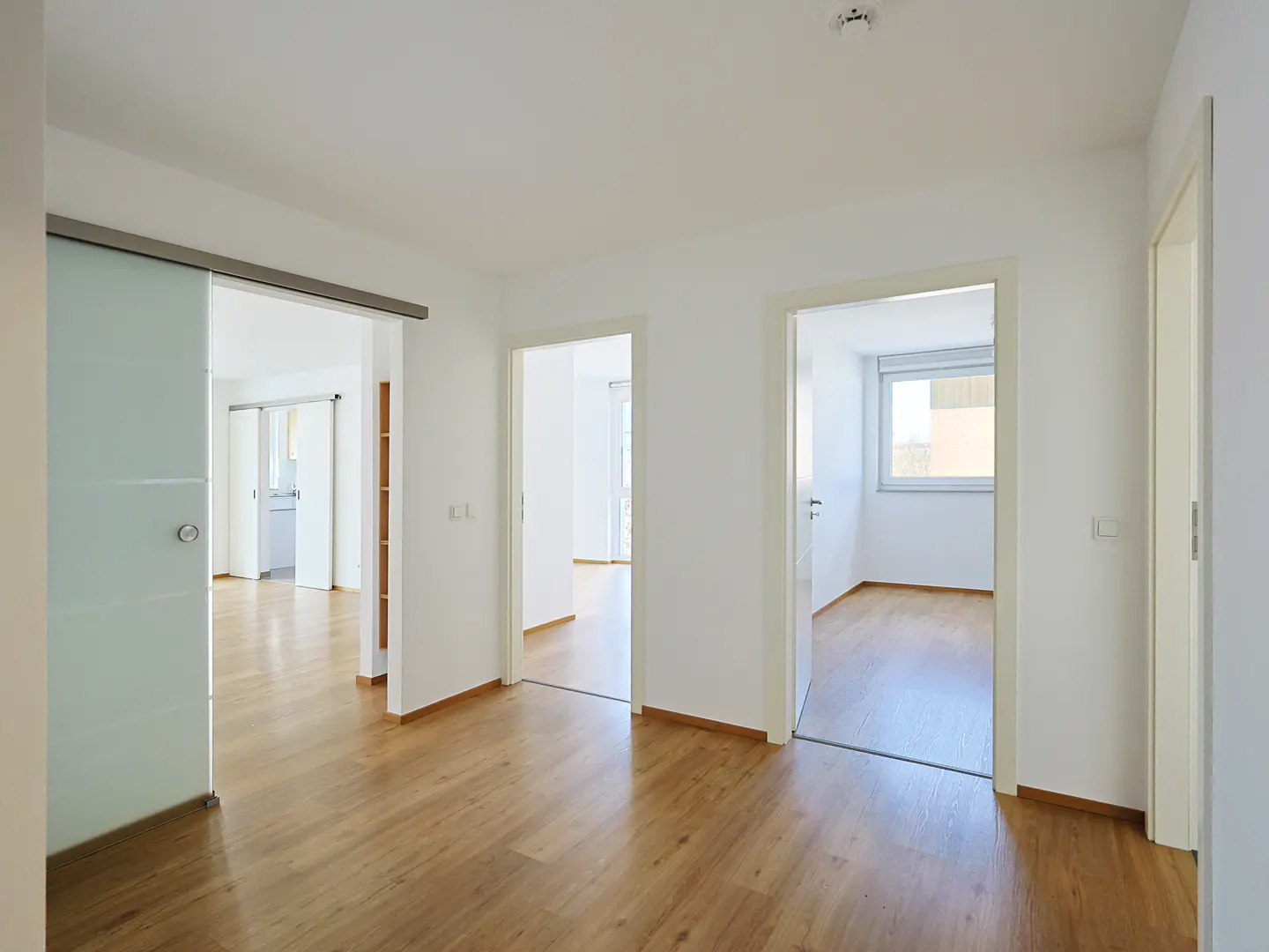 Bright, empty room with wood floors, white walls, and multiple doorways. A frosted glass sliding door is on the left.