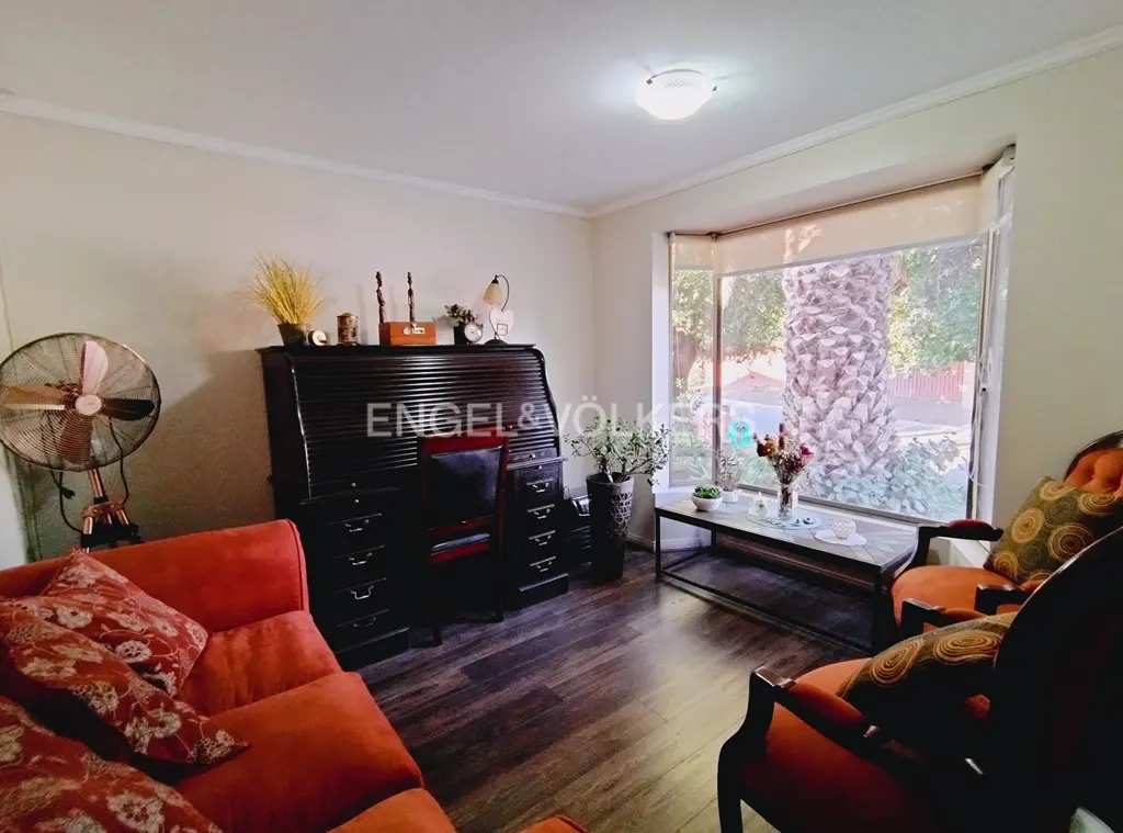 Living room with dark wood floors, orange sofa and chairs, and a roll-top desk under the Engel & Völkers logo. A large window shows a palm tree.