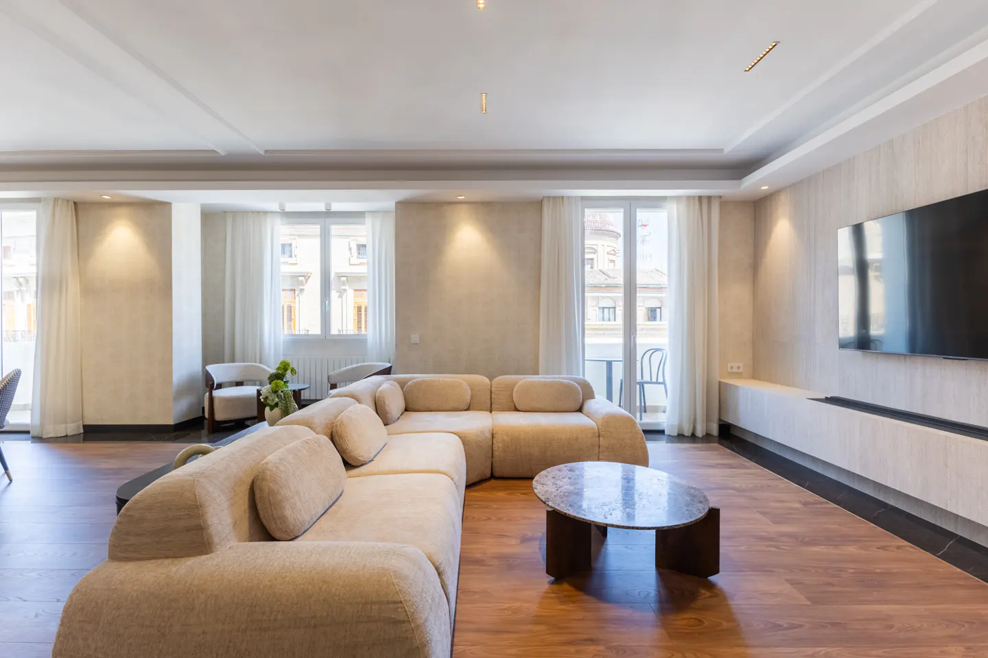 Modern living room with a beige sectional sofa, marble coffee table, wood floors, and a large wall-mounted television.