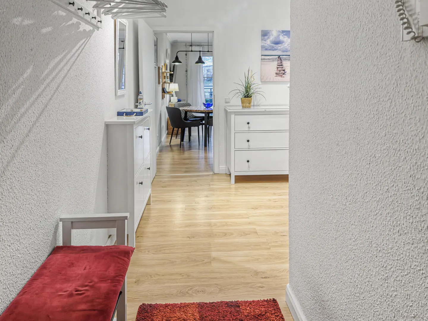 Bright hallway with white walls, light wood floors, a red bench, and a view into a dining area with a table and chairs.