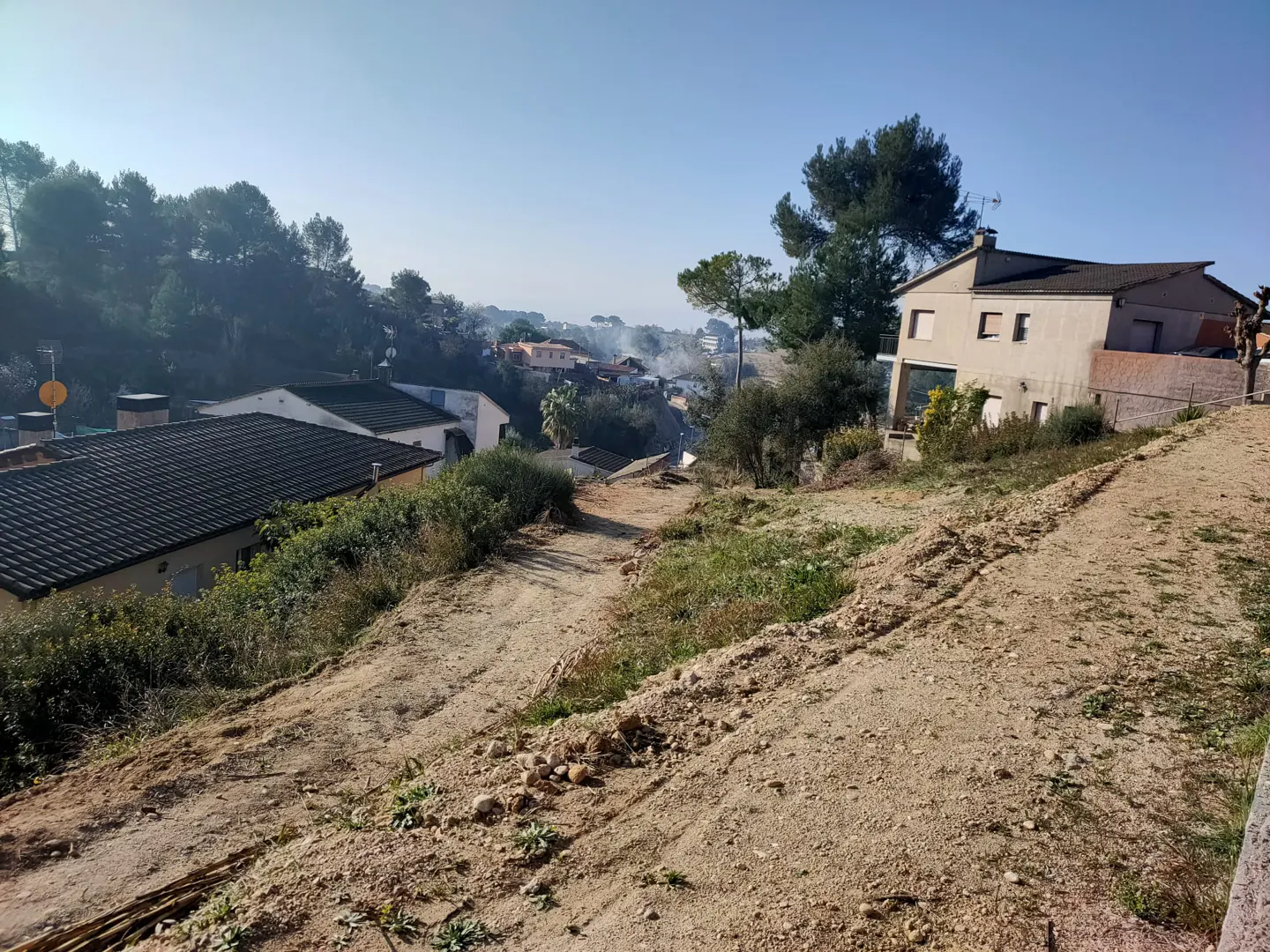 View of a vacant lot with a dirt path leading to houses in the distance, under a clear blue sky.
